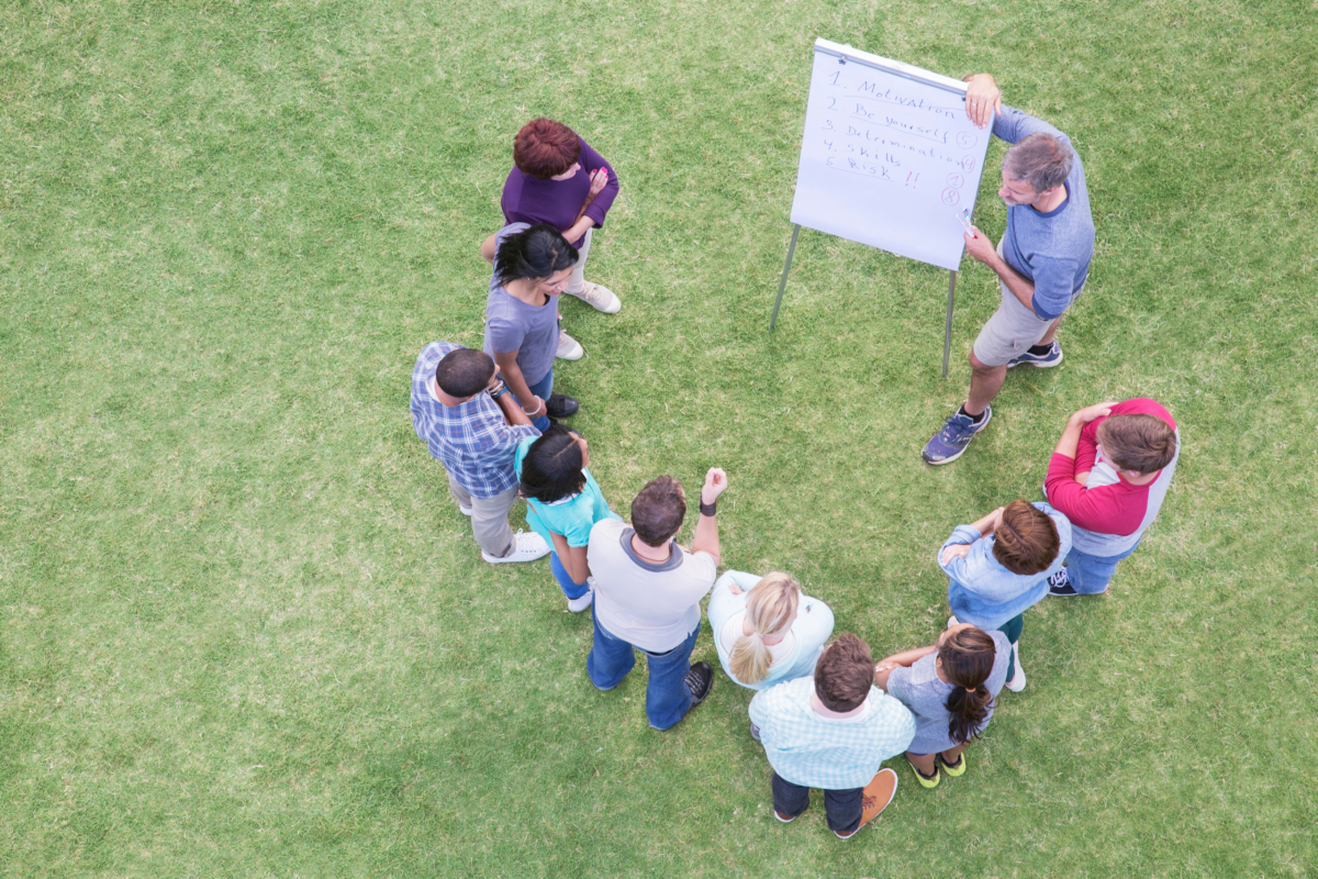 Eine Gruppe Menschen steht auf einer grünen Wiese vor einem Flip-Chart.