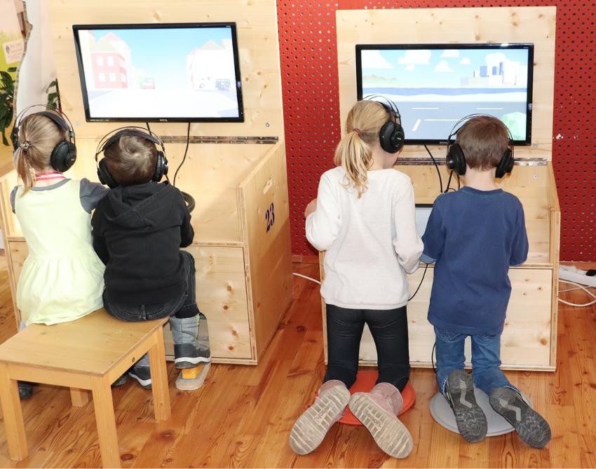4 Kinder sichtbar von hinten, sie knien und sitzen vor Holzstationen der Klimaversum-Wanderausstellung, und schauen auf Bildschirme.