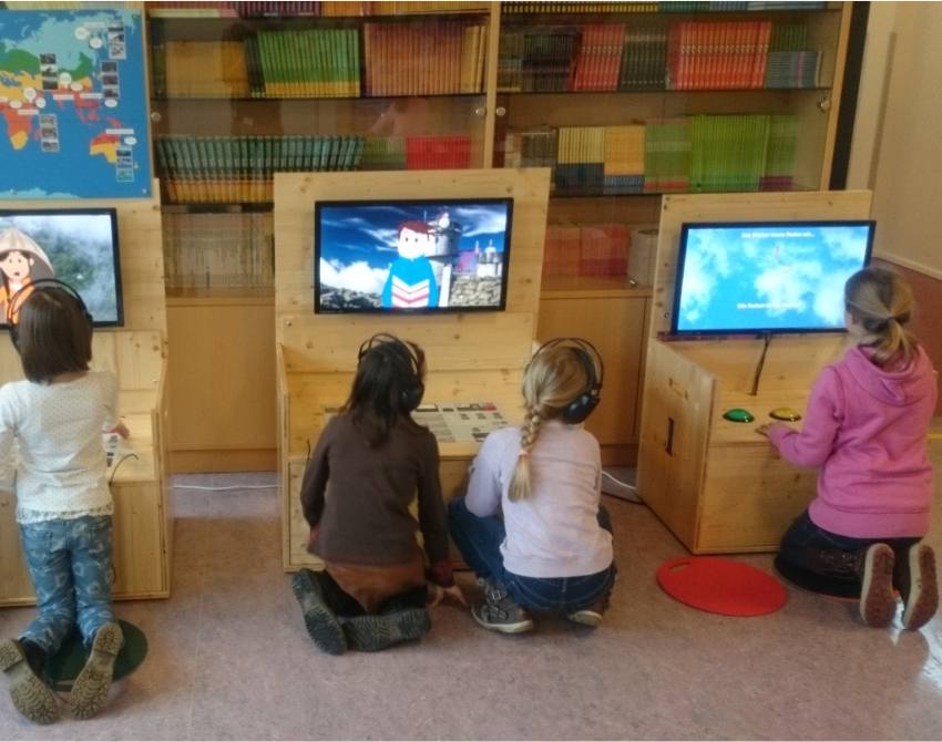 vier Kinder sitzen vor Stationen der KLIMAVERSUM Wanderausstellung vor Bildschirmen, mit dem Rücken zum:zur Betrachter:in