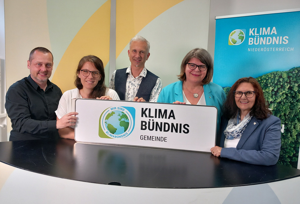 5 Menschen stehen hinter einem Stehpult und halten die "Klimabündnis-Gemeinde" Ortstafel.