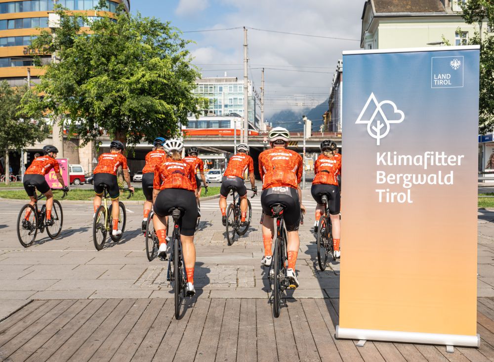 Ungefähr 10 Leute radeln in roter Radkleidung los, rechts steht ein "Klimafitter Bergwald Tirol" Banner