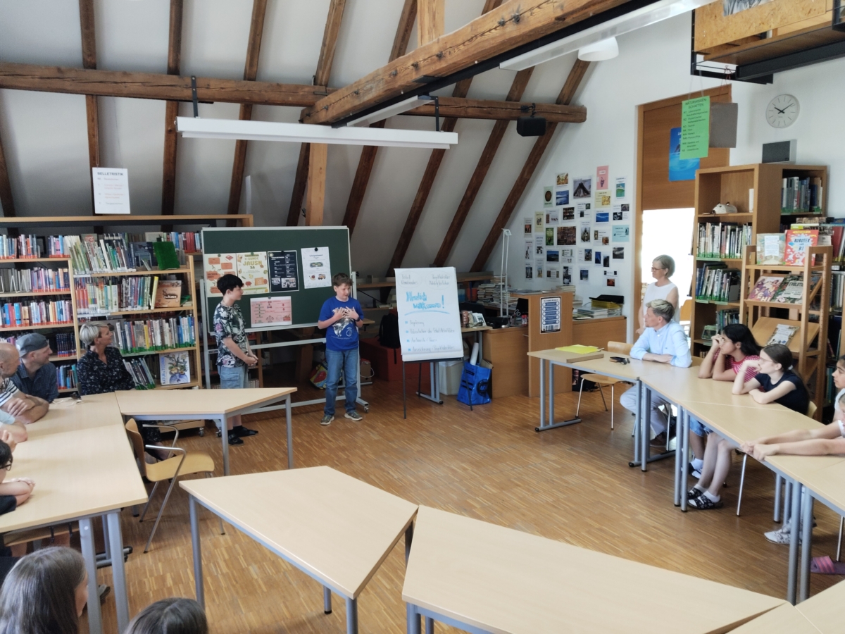 Zwei Schüler präsentieren an Tafel vor einem Klassenraum mit Jugendlichen und Erwachsenen