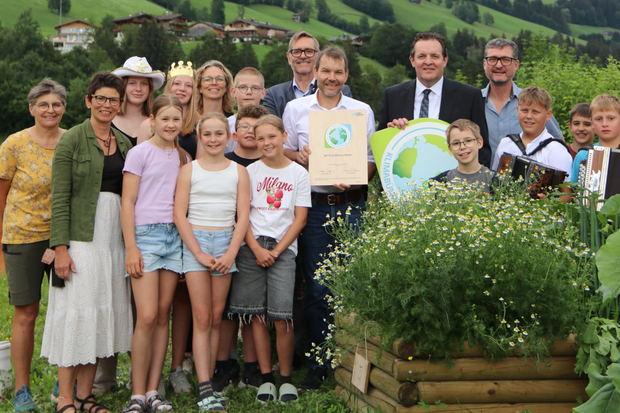 Fröhliche Kinder und Erwachsene im Grünen vor Wiesen und Bäumen mit Urkunden und Logo des Klimabündnis
