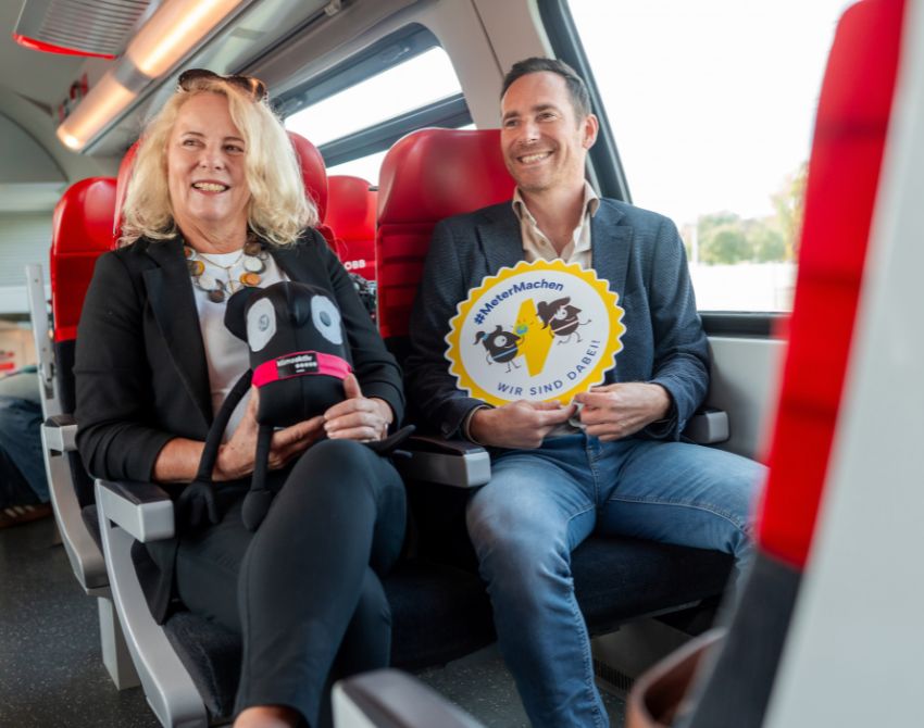 Frau mittleren Alters und Mann mittleren Alters fahren im Bus und halten Maskottchen der Europäischen Mobilitätswoche und ein Schild